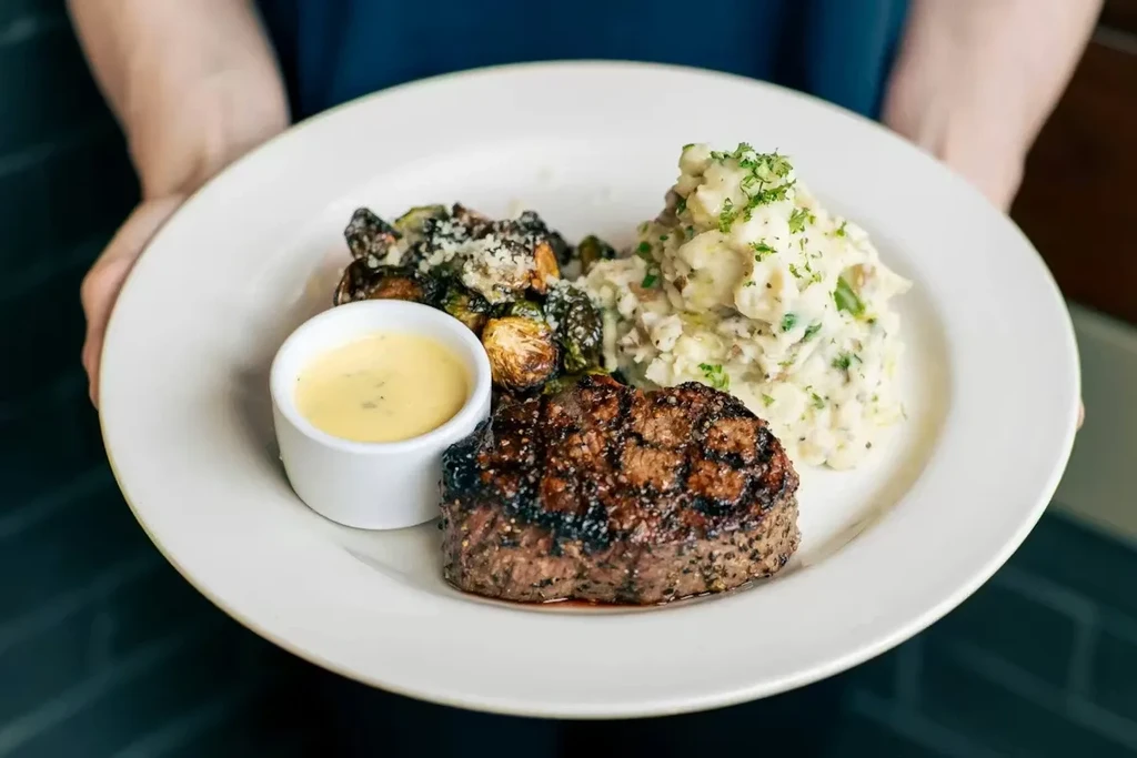 Hands holding plate with grilled steak, sauce, Brussels sprouts, mashed potatoes.