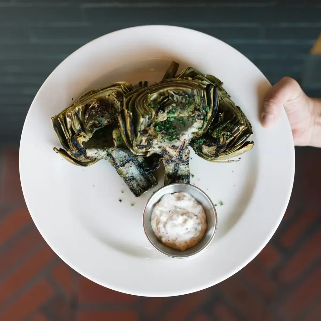 Hand serving grilled artichoke halves with herbs and dipping sauce.