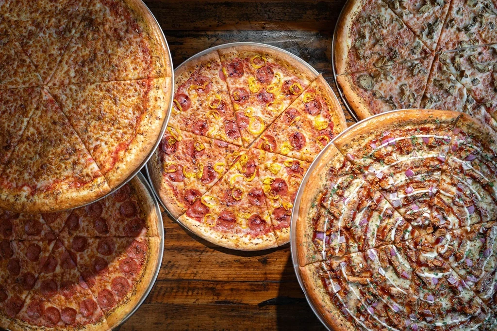 Five various pizzas on metal pans on a rustic wooden table.