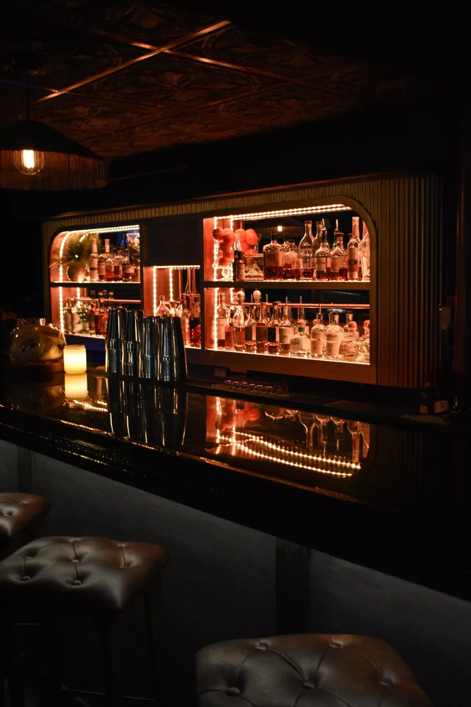 Illuminated liquor bottles on a dark bar counter with tufted stools.