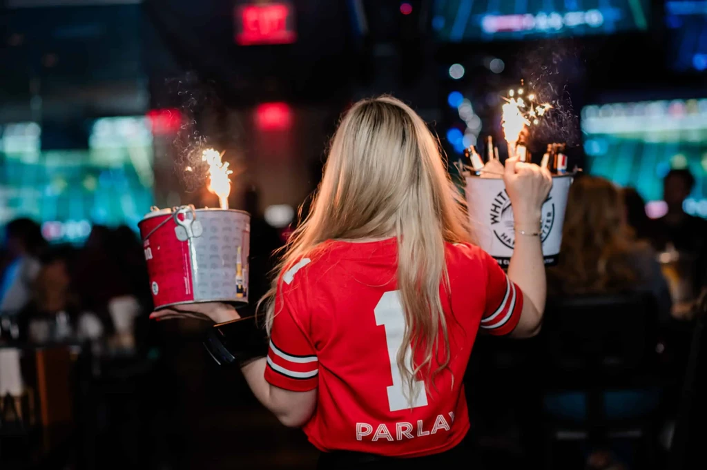 Person in '1 PARLAY' shirt holds two buckets with bright sparklers.