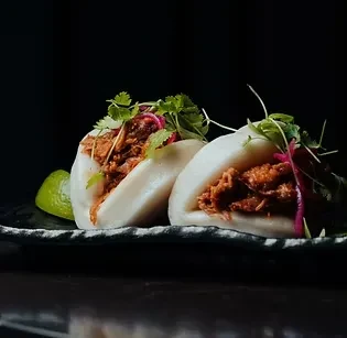 Two steamed bao buns with saucy shredded meat, cilantro, onions, lime.