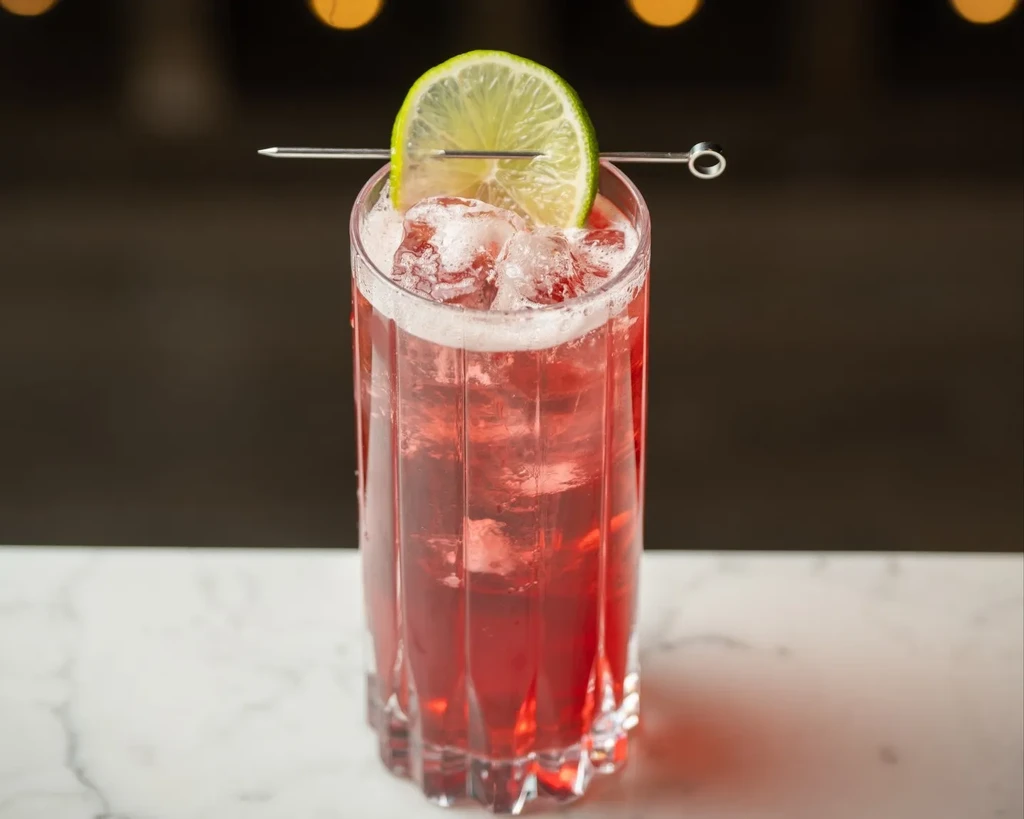 Ribbed glass with red cocktail, ice, foam, and lime garnish.