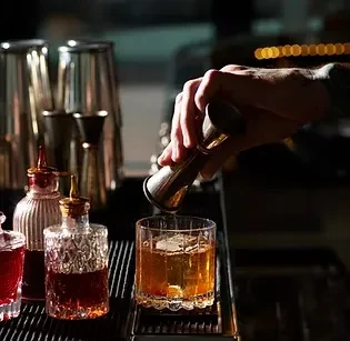 Hand pours from jigger into glass with ice and amber cocktail.