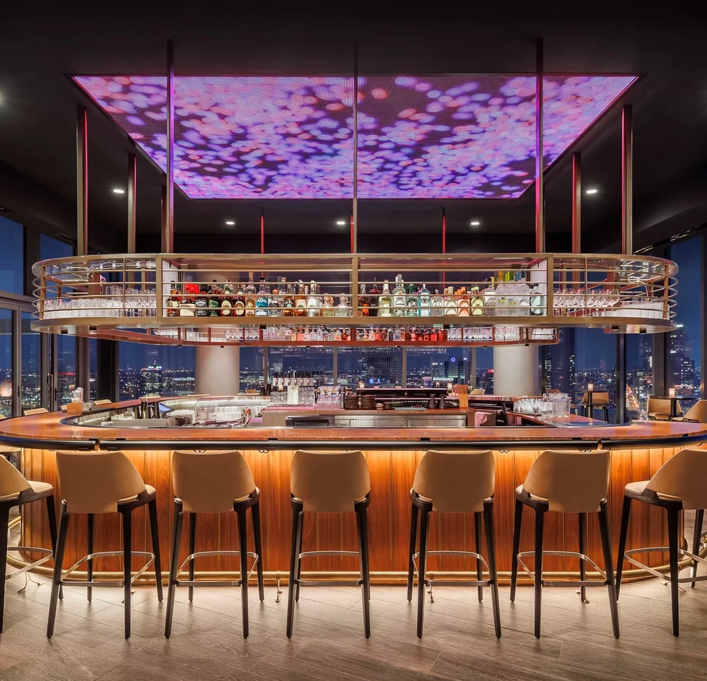 Modern bar with curved wood counter, abstract LED ceiling, and city skyline.