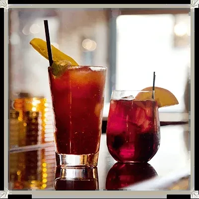 Two garnished cocktails, reddish-brown and dark red, reflecting on a bar.