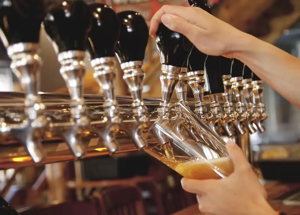 Hands pour golden beer from a chrome tap into a clear glass.