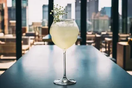 Tall cocktail with light drink, ice, white flowers, blurred city view.
