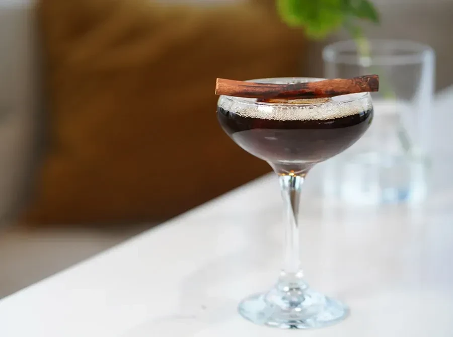 Dark cocktail in coupe glass with foam and cinnamon stick.