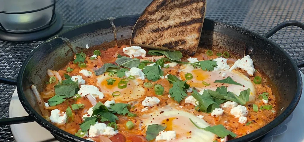 Shakshuka with eggs, cheese, herbs, and bread in a cast iron pan.