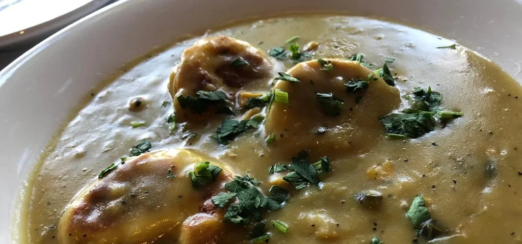 Creamy soup with golden dumplings and green herbs in a white bowl.