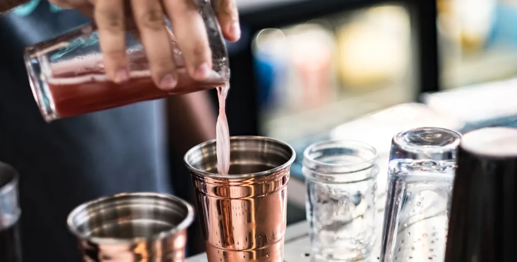 Close-up of hand pouring pink liquid into a copper cocktail shaker.