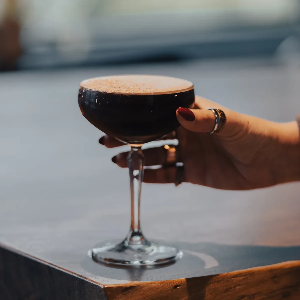 Red-nailed hand holds a foamy dark cocktail in a delicate coupe glass.