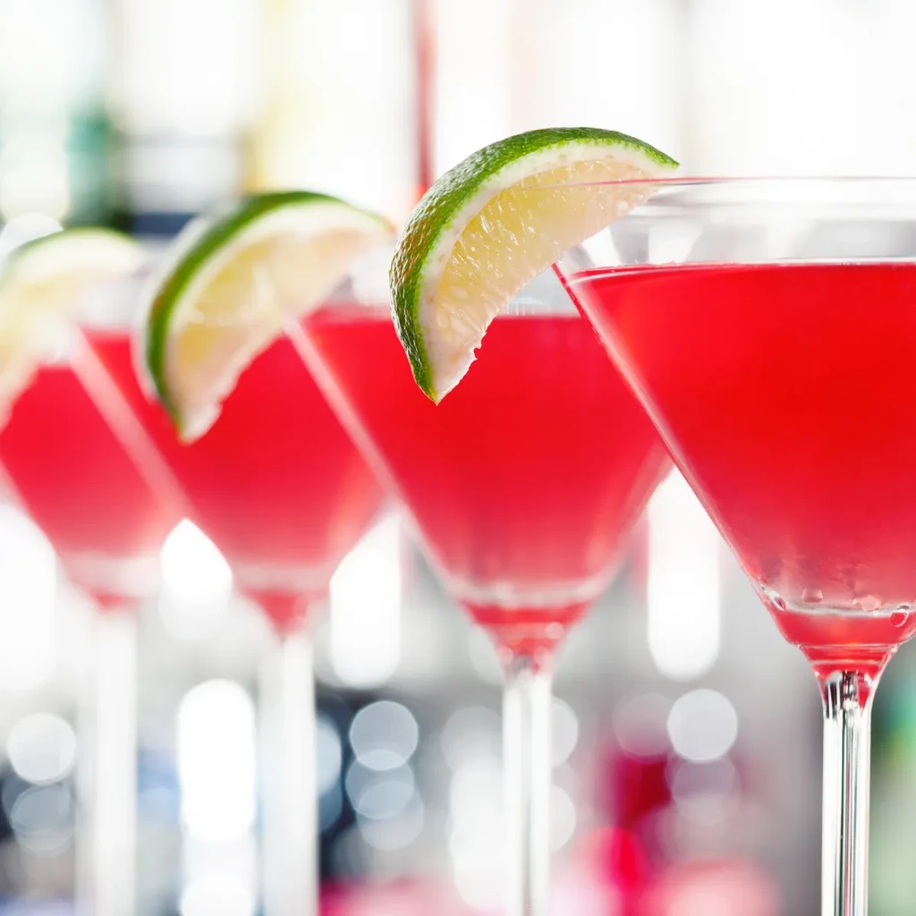 Close-up of four red cocktails with lime, blurred bar background.