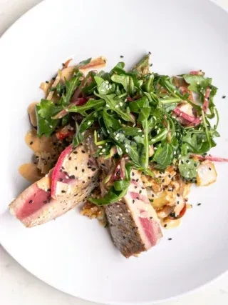 Seared sesame tuna slices, arugula salad, and creamy sauce on a plate.