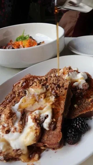 French toast with cream, nuts, blackberries, and syrup being poured.