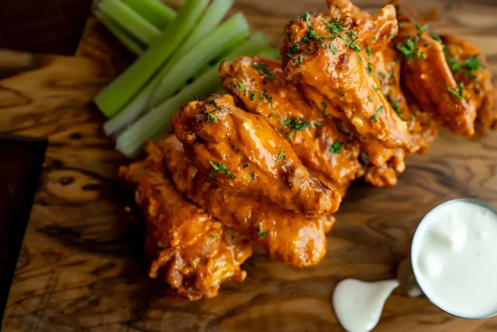 Buffalo chicken wings, celery sticks, and dip on a wooden board.