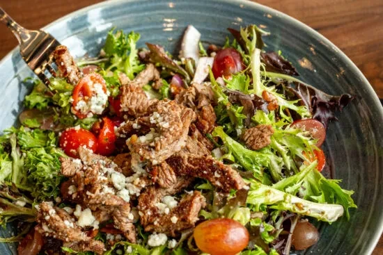 Close-up of steak salad with greens, tomatoes, cheese, and grapes.