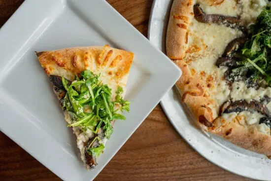 Overhead view of pizza slice, full pizza, arugula, on wooden table.