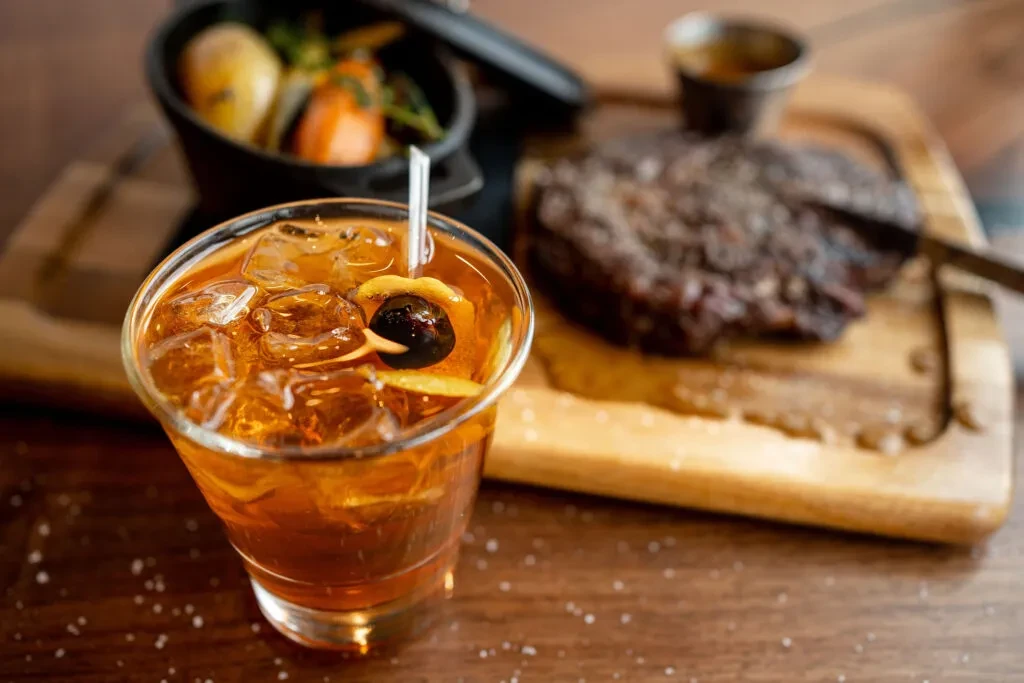 Garnished amber cocktail, seasoned steak, and vegetables on a dark wooden table.