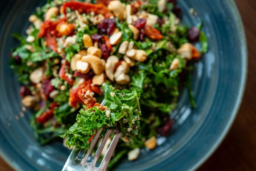 Fork lifts kale from salad with tomatoes, cashews in blue bowl.