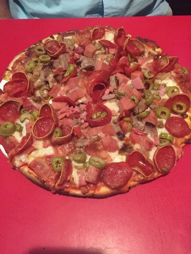 Pizza with pepperoni, ham, sausage, olives, green peppers on a red table.