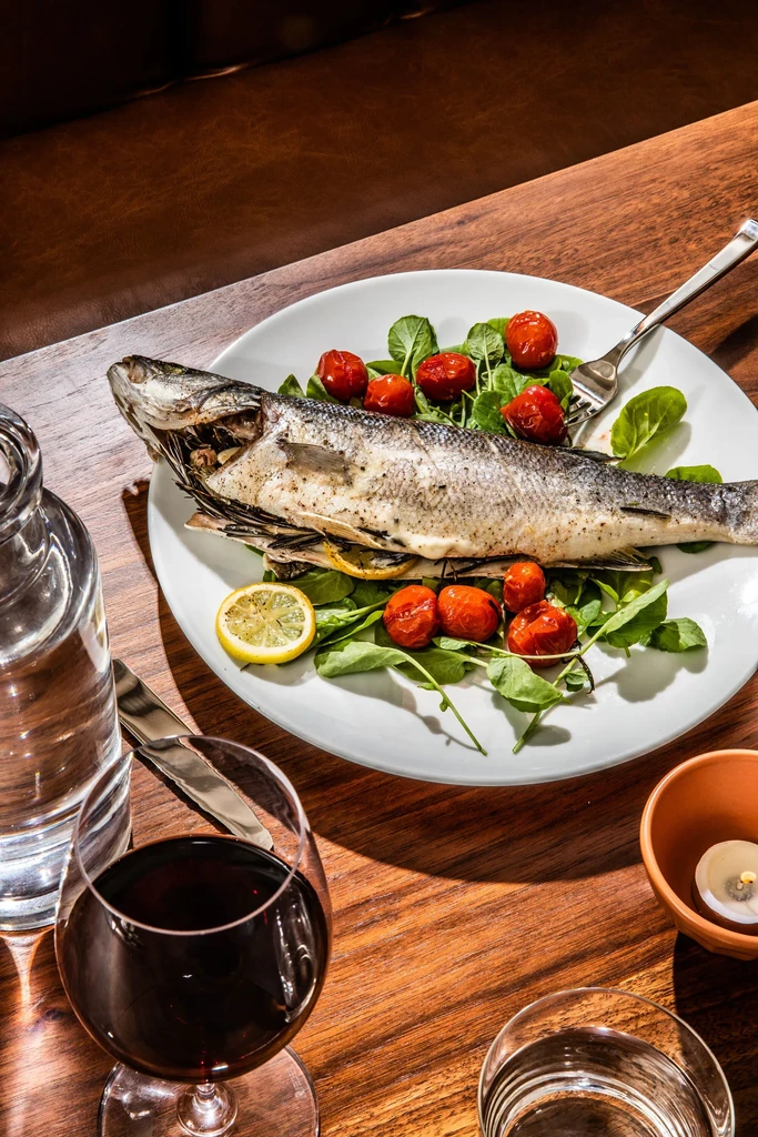 Roasted fish, greens, tomatoes, lemon, wine, candle on dark wood table.