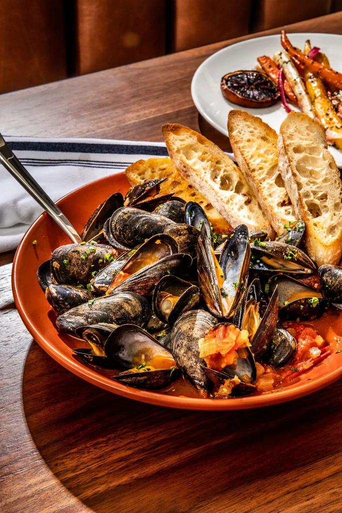 Steamed mussels, bread, roasted vegetables, and lemon on a wooden table.