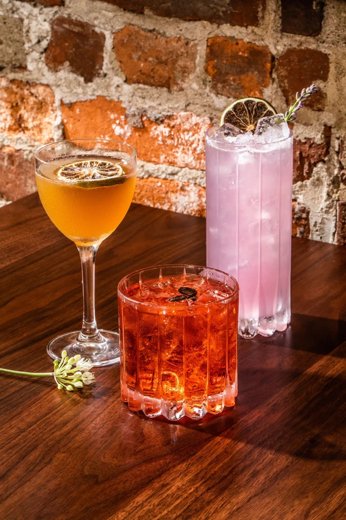 Three distinct garnished cocktails on a dark table with a brick wall.