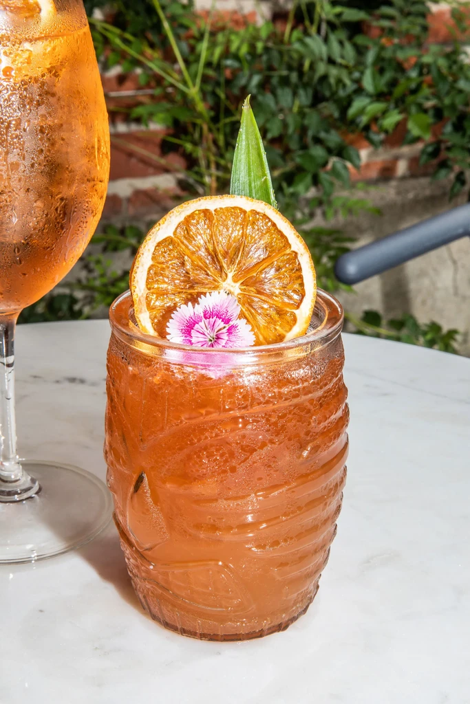 Heavily condensed orange tiki cocktail with elaborate garnish and second drink.