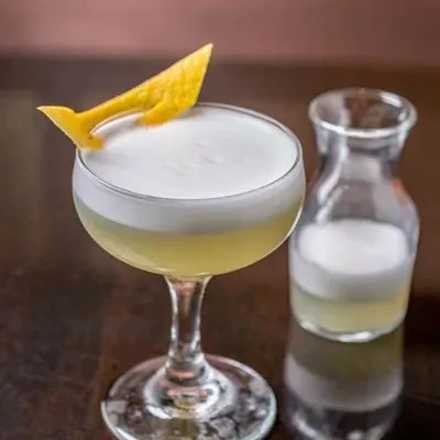 Coupe glass cocktail with white foam, lemon twist, and small carafe.