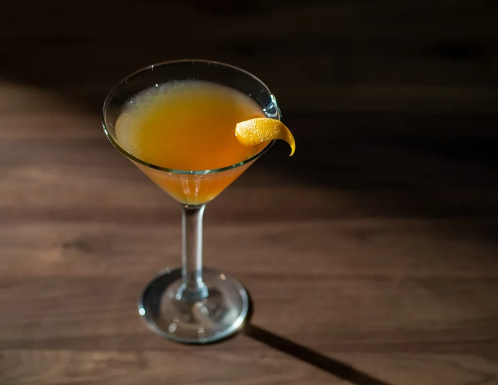 Dramatically lit orange cocktail in martini glass with orange peel on wood.
