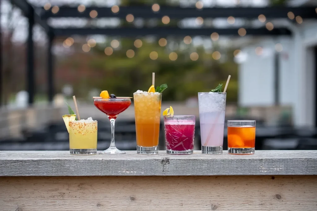 Six diverse cocktails on a rustic bar with outdoor string lights.