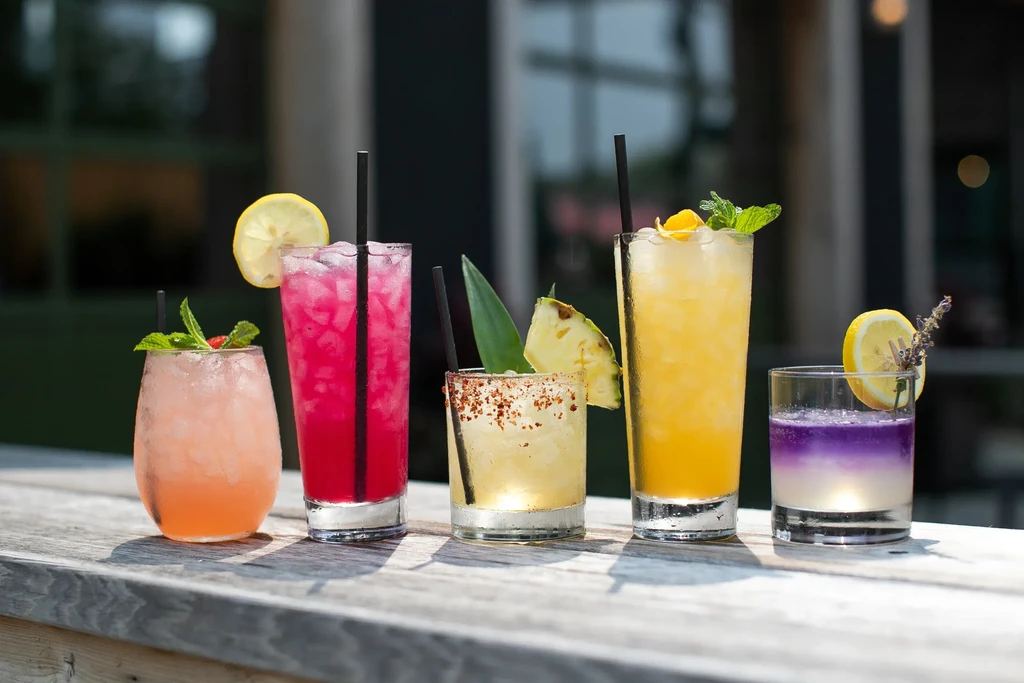Five brightly colored cocktails with garnishes and straws on a wooden surface.