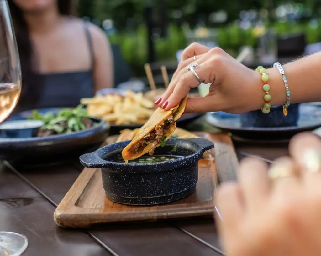 Hand with red nails dipping a golden meat taco into a sauce bowl.