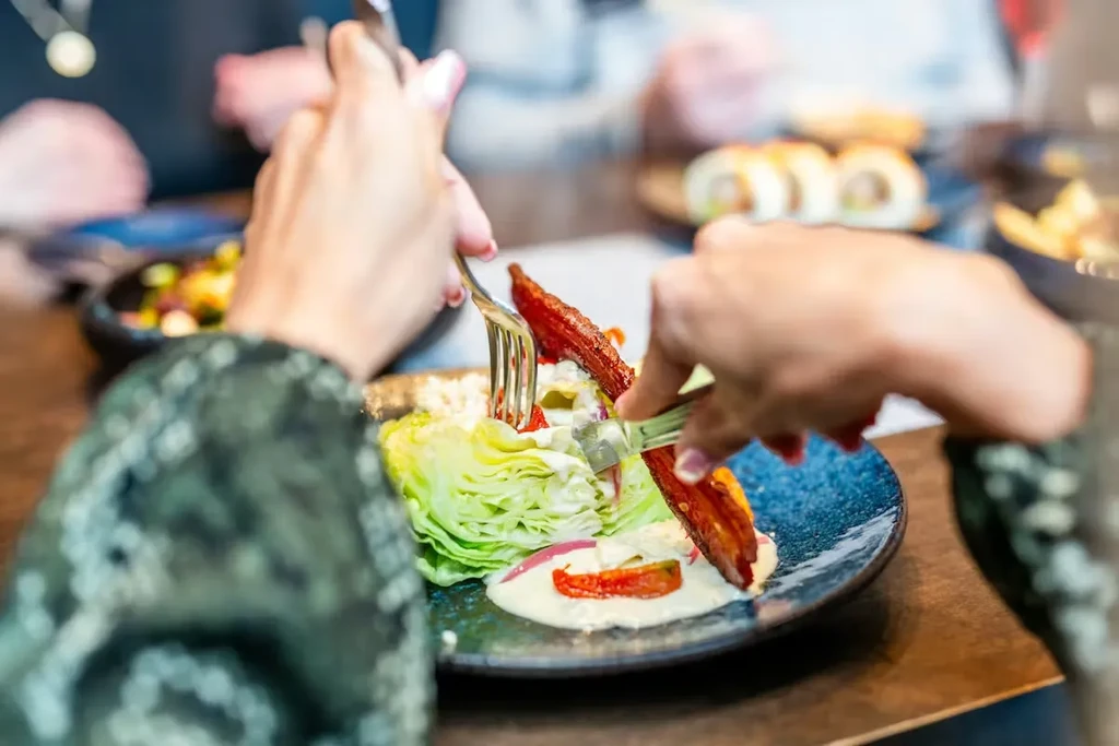 Hands cutting a wedge salad topped with bacon, dressing, and red onion.
