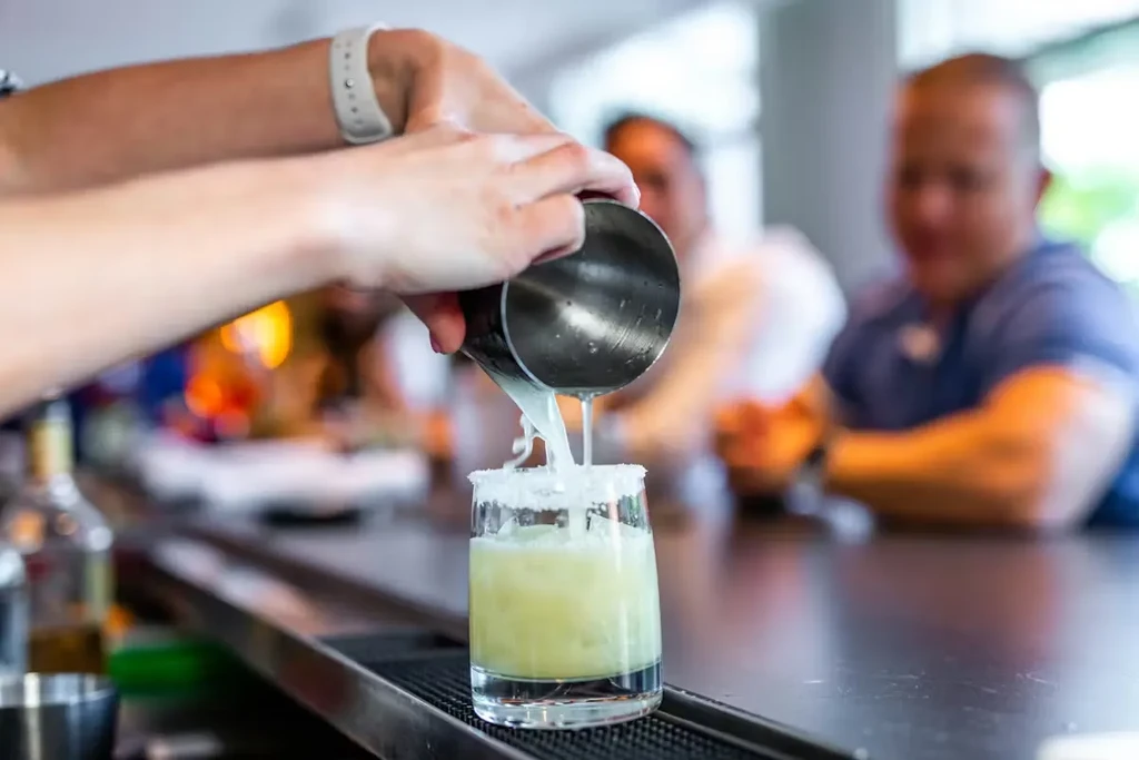 Bartender's hands pouring green drink from shaker into salt-rimmed glass.