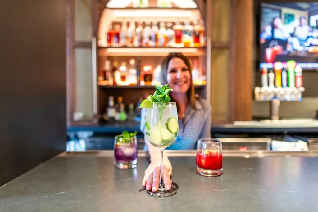 Three colorful cocktails on a bar, with a smiling bartender in soft focus.