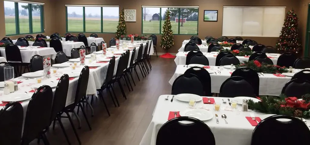 Festive dining hall with decorated tables, Christmas trees, and rural view.