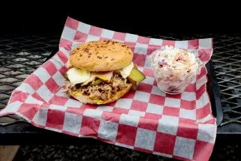 Pulled meat sandwich with pickles and coleslaw on checkered paper.