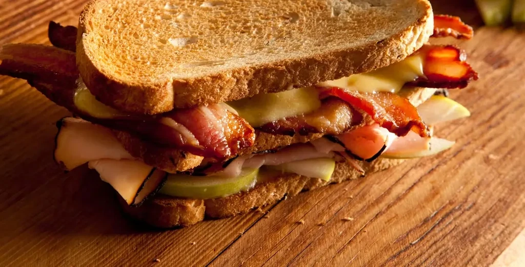Toasted club sandwich with bacon, melted cheese, green apple, on wood.