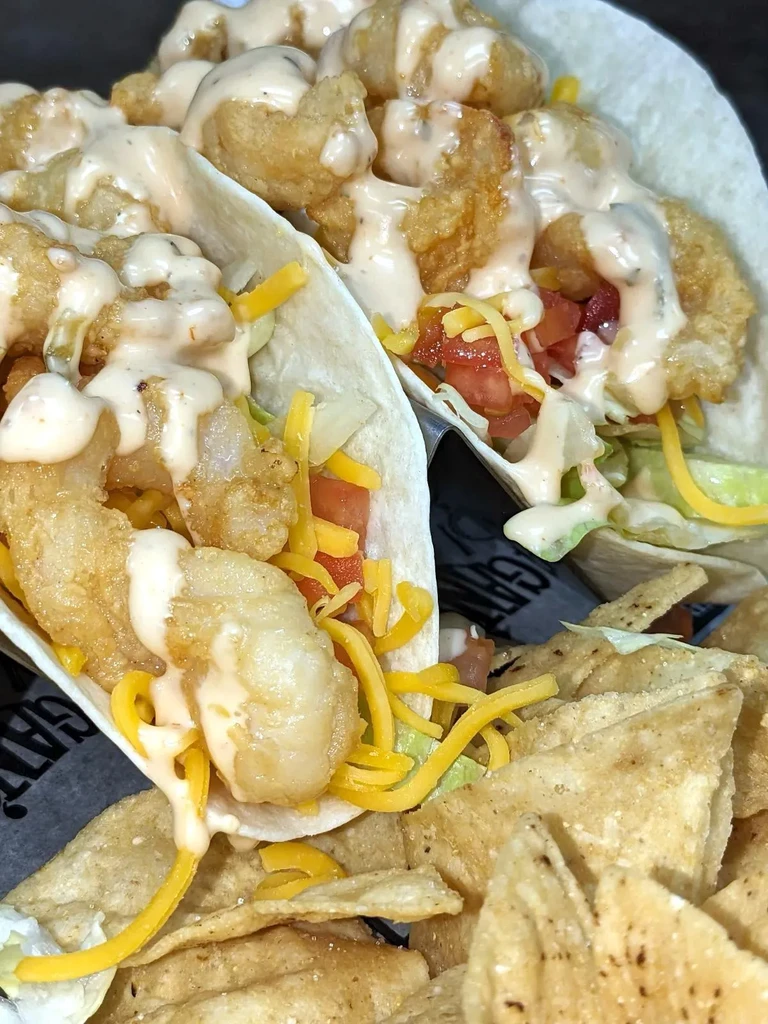 Two crispy fried shrimp tacos with lettuce, tomato, cheese, sauce, and chips.