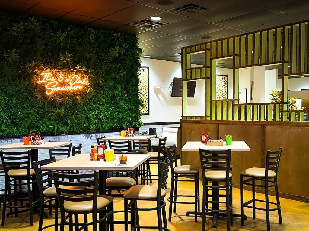 Restaurant dining area with high-top tables and neon sign on plant wall.