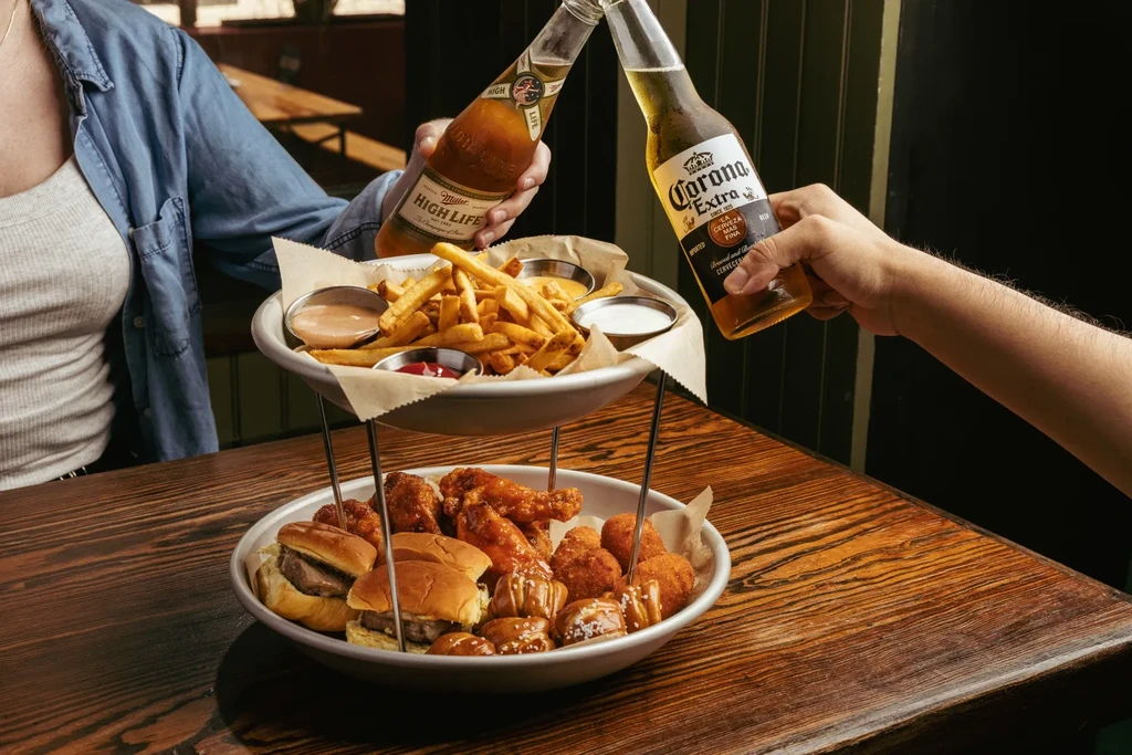 Two people toast beer bottles above tiered snacks on a table.