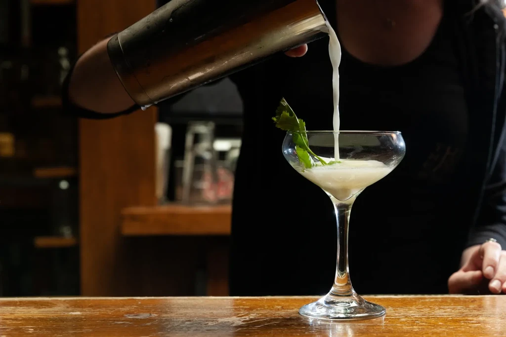 A bartender pours a light-colored drink from a shaker into a coupe glass.