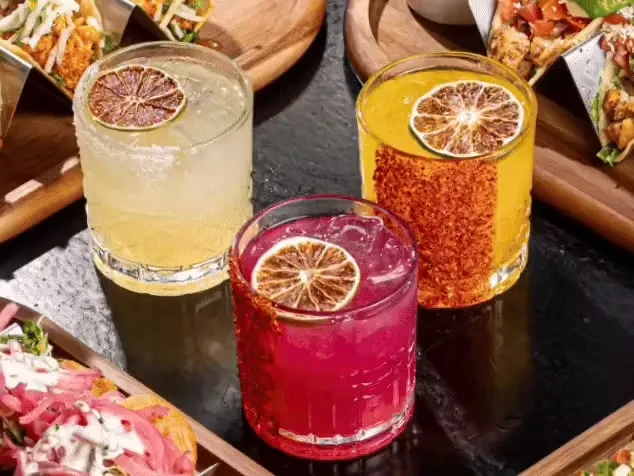 Three colorful cocktails garnished with dried lime slices, next to tacos.