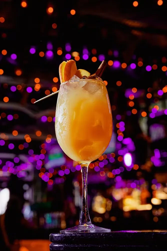 A golden cocktail in a wine glass with cinnamon and fruit garnish.