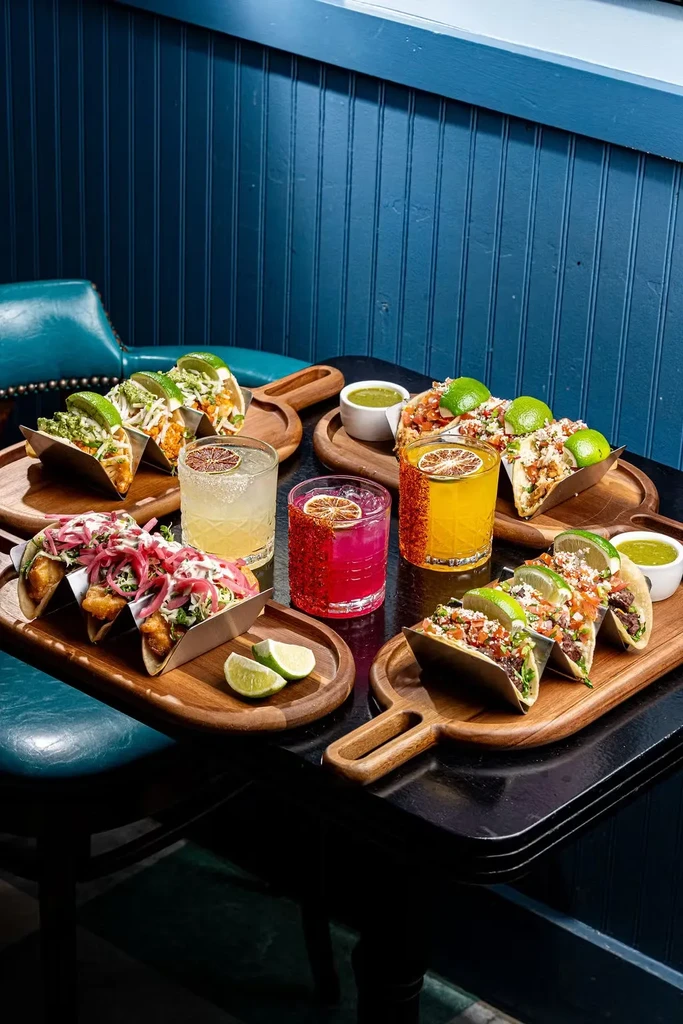 Multiple tacos, drinks, and lime wedges on wooden serving boards.