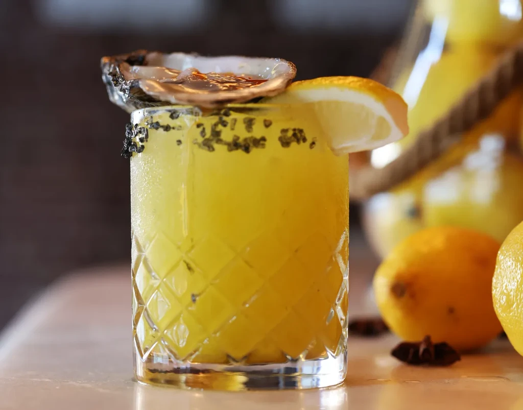 Yellow cocktail with an oyster and lemon wedge garnish.