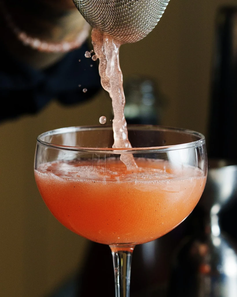 A pinkish-orange drink is poured from a strainer into a coupe glass.